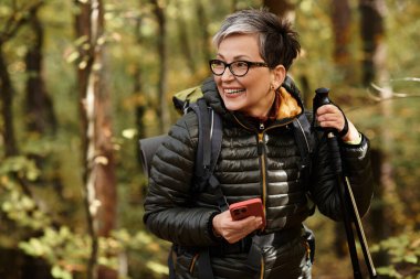 Son sınıf öğrencisi bir kadın elinde telefon ve yürüyüş direğiyle renkli bir sonbahar ormanında yürüyüş yaparken gülümsüyor..