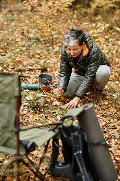A senior woman kneels on the forest floor, cooking a meal amid autumn leaves and natures beauty.