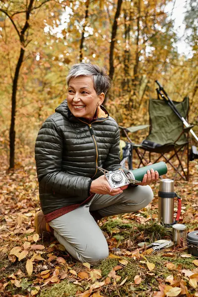 Son sınıf öğrencisi bir kadın, canlı sonbahar yaprakları üzerinde diz çöker, doğa yürüyüşüne hazırlanırken gülümser..