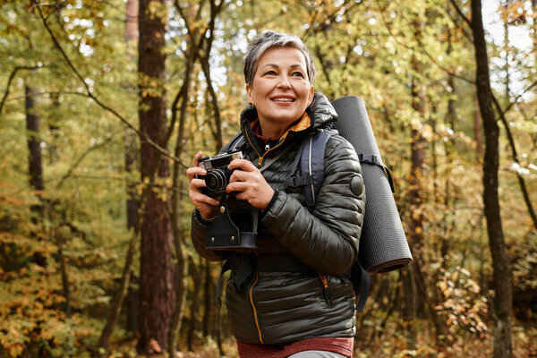 Senior woman enjoys hiking in a colorful forest, capturing memories with her camera.
