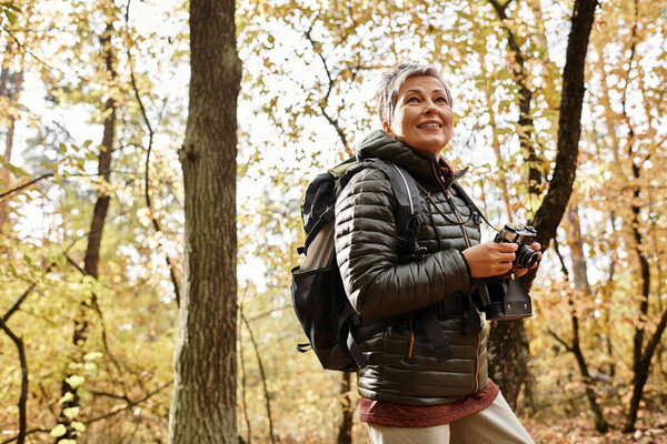 Senior woman enjoys the beauty of a colorful forest while capturing memories with her camera.