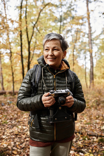 A senior woman smiles warmly while hiking in a beautiful autumn forest, holding her camera.