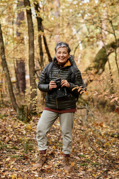 A happy senior woman pauses to capture the beauty of a colorful autumn forest while hiking.