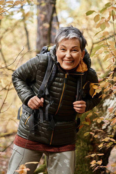 A senior woman with a bright smile enjoys her hike through a vibrant autumn forest.