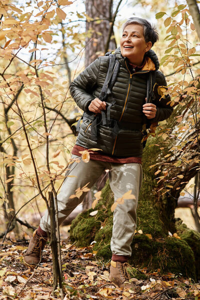 A senior woman enjoys hiking in a vibrant autumn forest, surrounded by colorful foliage.