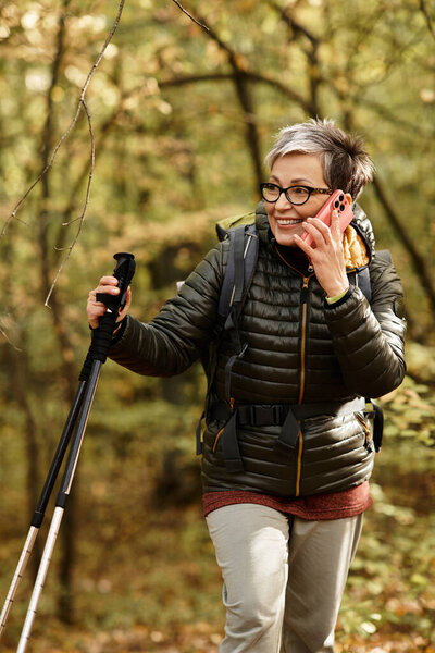 A senior woman traverses a lush forest trail, smiling and talking on her phone with excitement.