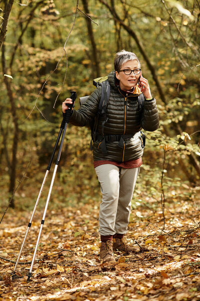 A senior woman is hiking on a forest trail surrounded by colorful autumn leaves, smiling.
