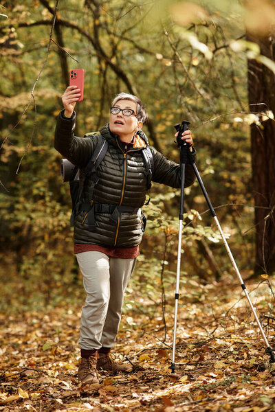 A senior woman with hiking gear explores a colorful forest, capturing the beauty of her journey.