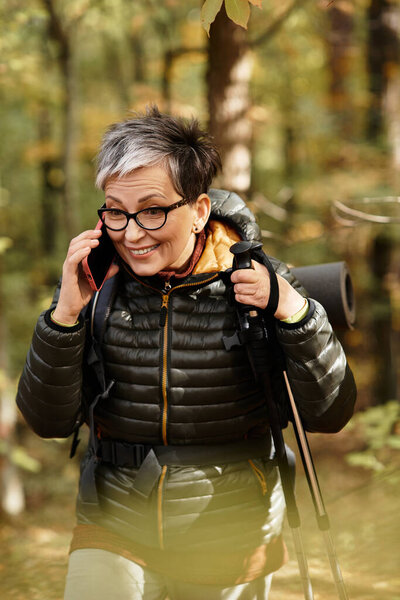 A senior woman in a cozy jacket hikes a forest trail while talking on her phone.