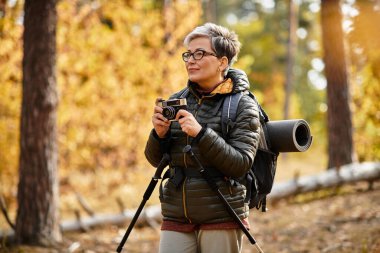 Güneşli bir ormanda, yaşlı bir kadın yürüyüşü sırasında büyüleyici sonbahar manzarasını yakalamak için durur..