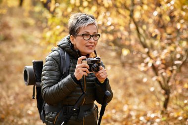 Kıdemli bir kadın dingin bir ormanı keşfediyor, heyecanlı bir sonbahar manzarasının fotoğraflarını çekiyor..