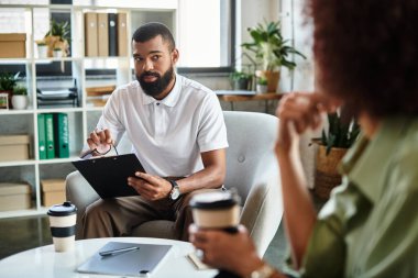 Genç bir adam, modern bir stüdyoda gün içinde bitkilerle ve doğal ışıkla bir kadınla röportaj yapıyor..