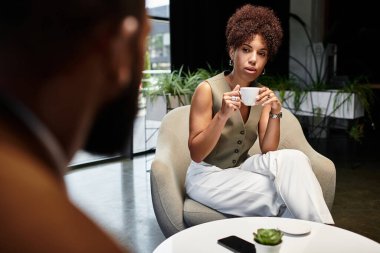 Genç bir kadın, bir röportaj sırasında modern bir stüdyoda içkisini yudumlarken dikkatle dinliyor..