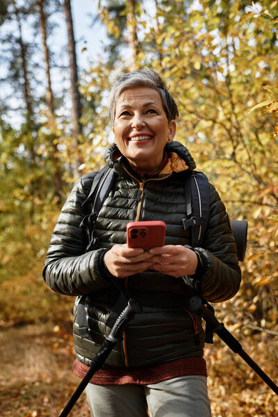 Senior woman enjoys hiking through a colorful forest while checking her phone with a smile.
