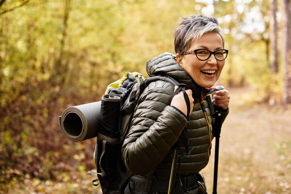 Senior woman smiles brightly while hiking through a vibrant forest path surrounded by autumn leaves.