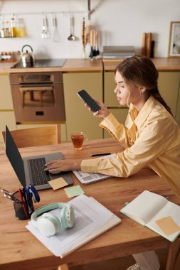 Evde dizüstü bilgisayar, akıllı telefon ve çayını yudumlayan genç bir kadın..