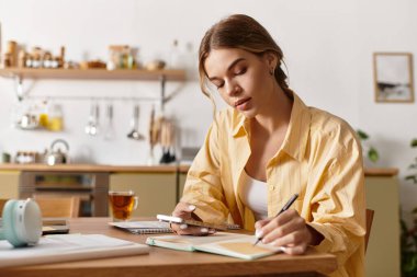 Yazmaya odaklanmış genç bir kadın ve evde rahat bir mutfak ortamında telefonunu kullanıyor.