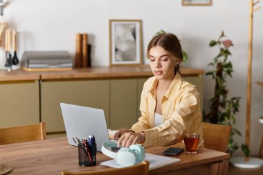 Genç ve güzel bir kadın, çay yudumlarken dizüstü bilgisayarına odaklanmış bir şekilde evdeki masasında oturuyor..