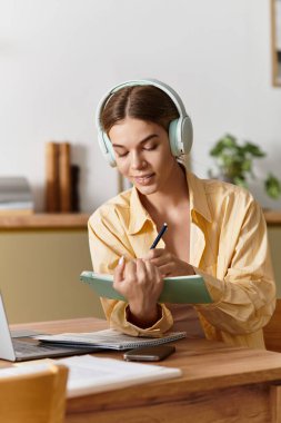 Genç bir kadın kitap dolu sevimli masasında notlar yazarken kulaklık takıyor..