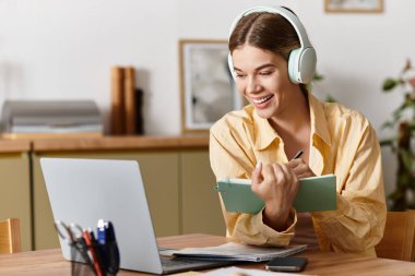 Genç bir kadın evde online öğrenim görüyor, notlar alırken gülümsüyor ve kulaklık kullanıyor..