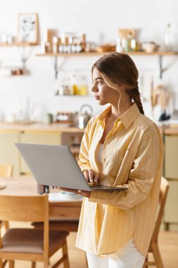 Genç ve güzel bir kadın, güneş içeri girerken rahat mutfağında dizüstü bilgisayarına odaklanmış duruyor..