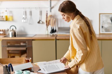 Güzel genç bir kadın işine odaklanmış, modern mutfağındaki notlara göz atıyor..