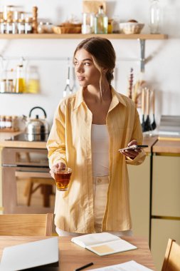 Genç bir kadın mutfakta dinleniyor, çayını yudumluyor ve telefonunu kullanıyor..