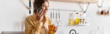 Genç ve güzel bir kadın çay yudumlarken ve mutfakta telefonla konuşurken gülümsüyor..