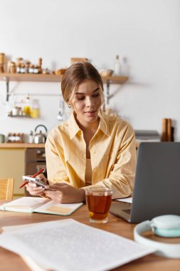 Günlük kıyafetlerini giymiş genç bir kadın mutfak masasında çayını yudumlarken görevlerini yapıyor..