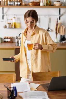 Sıcak bir mutfakta, çayın tadını çıkaran ve telefonunu karıştıran genç bir kadın rahatlatıcı bir atmosfer yaratıyor..