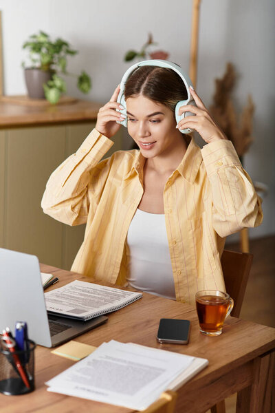 A young woman wearing headphones is focused on her studies at home, enjoying a cup of tea.
