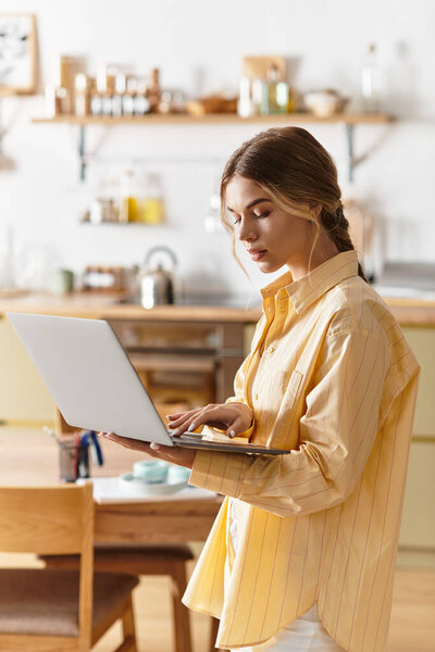 In a bright kitchen, a beautiful young woman focuses on her laptop, immersed in her work.