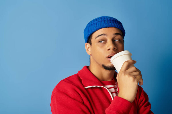 A stylish young man wearing a blue beanie casually sips coffee while posing against a blue wall.