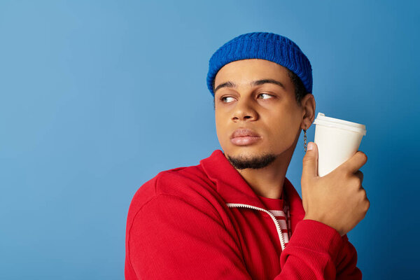 Stylishly dressed young man enjoys his drink while showcasing vibrant fashion on a blue wall.