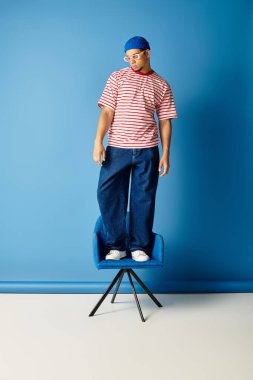 Modaya uygun genç bir siyah adam mavi bir sandalyede poz veriyor, mavi bir duvara karşı modaya uygun bakışını vurguluyor..