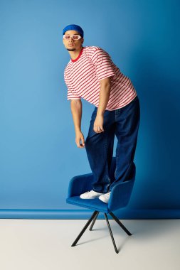 Modaya uygun genç bir Afrikalı Amerikalı adam mavi bir sandalyede kendine güvenerek duruyor, şık bir şekilde poz veriyor..