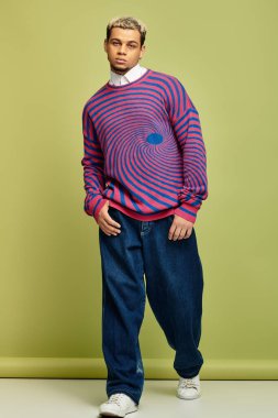 Soyut çizgili kazak giyen şık bir genç adam yeşil bir arkaplanda güvenle poz veriyor..