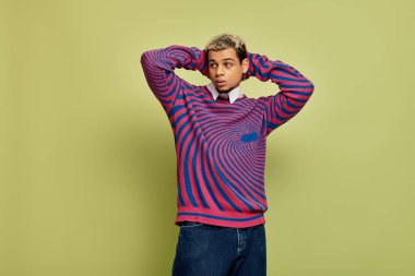 Tarz sahibi genç bir adam kendine güvenerek yeşil renklerin arasında çizgili bir kazak giyiyor..