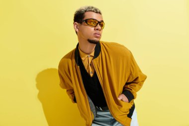 Genç bir siyah adam sarı bir arka plana karşı şık bir bombacı ceketiyle güven içinde poz veriyor..