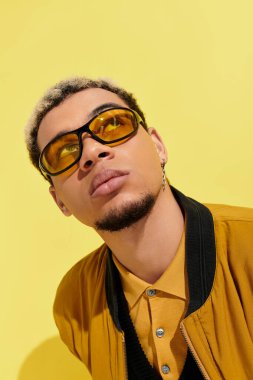 Şık bir genç adam parlak sarı bir stüdyoda bombacı ceketi ve güneş gözlüğü modelliyor..