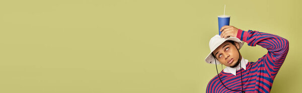 Стильный молодой чернокожий мужчина игриво позирует с напитком на голове в абстрактной толстовке, баннер