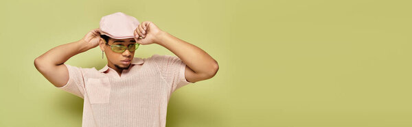 Стильный молодой чернокожий мужчина моделирует розовое поло и плоскую кепку в ярко-зеленой обстановке, баннер