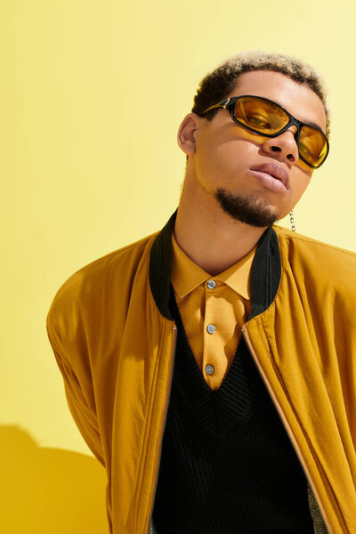 Молодой черный человек в стильной куртке бомбардировщика уверенно позирует на ярко-желтом фоне.