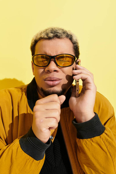 A young Black man in a stylish bomber jacket talks on his phone against a bright yellow background.