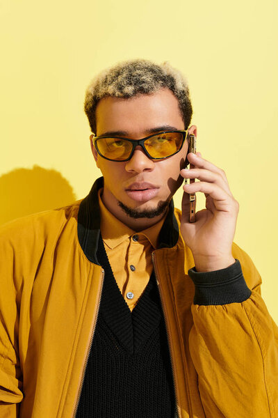 A young Black man shows off his style on a phone call against a bright yellow background.