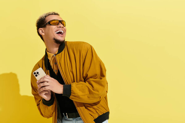 A trendy young man smiles joyfully while holding a smartphone, dressed in a stylish bomber jacket.