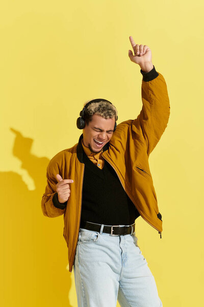 Молодой человек радостно танцует в студии в гладкой желтой куртке бомбардировщика и наушниках.