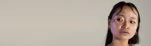 A young Asian woman showcases unique makeup artistry with pearls adorning her face in a studio, banner