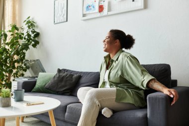 Rahatlamış bir kadın evdeki modern bir kanepede gülümsüyor, yeşilliklerle çevrili..