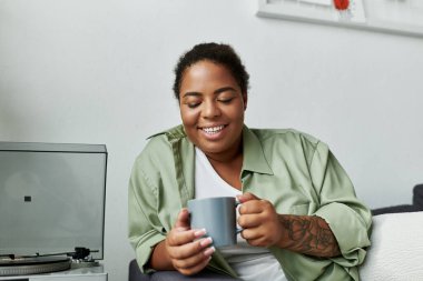 Modern bir dairede dinlenen bir kadın, sıcak gülümseyerek kahvesinin tadını çıkarır..